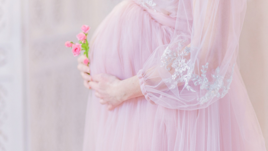 Beautiful pregnant woman in rich pink dress holds hands on her belly posing in luxury white room